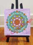 photograph of a square painting on paper, purple background with mandala primarily in blue, green, red, on a small black easel on a wood floor