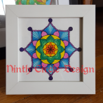Photograph of a mandala, a 8-fold symmetric pattern painted in mostly blues, purples, and greens with a white background, in a white frame