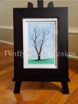 photograph of a framed painting/drawing of a leafless tree (drawn) against a background of blue sky and green grass (painted)