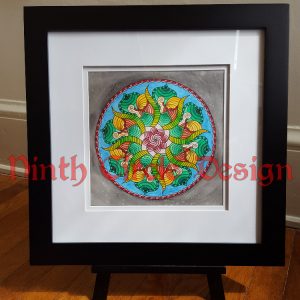 photograph of a mandala in greens and reds within a blue circle and grey background, framed in a black frame, white mat