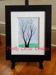 photo of a framed painting with blue sky, green grass, and the silhouette of a leafless tree; frame is black with white mat